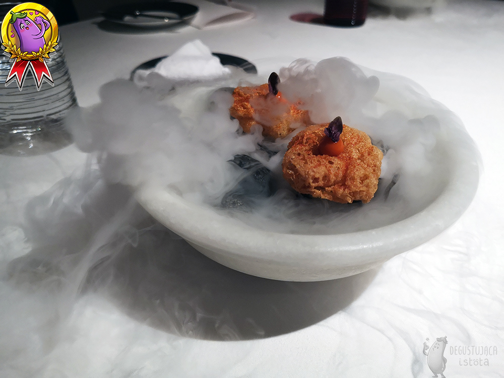 A white bowl from which vapor clouds of liquid nitrogen escape. In the middle you can see two crispy, orange flat cakes. On each cake there was a ball of orange gel with a brown leaf.