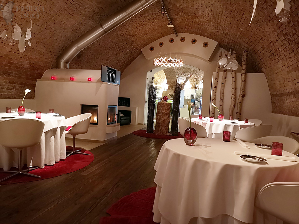 The hall is located in the basement lined with red bricks. Round, large tables are covered with white tablecloths. Each table is placed on a round, red carpet. On the tables are red glasses and a vase with a white calla.
