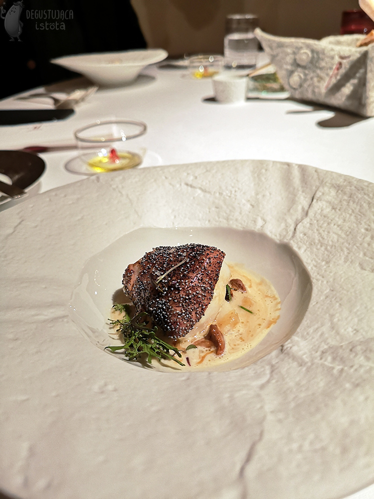 In a white, flat plate, the texture of which resembles marble, lies a portion of fish coated in poppy seeds. Under the fish you can see orange and white sauce and pieces of chanterelles.