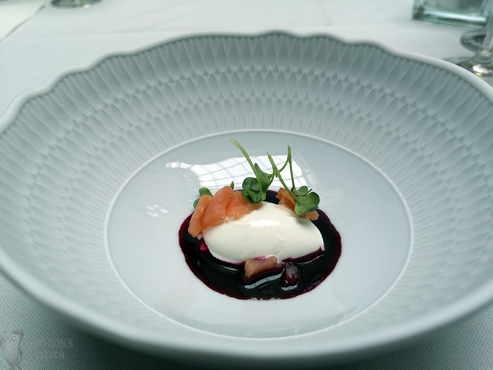 On a blue plate lies a portion of white ice cream surrounded by beet consomme and garnished with pieces of salmon.