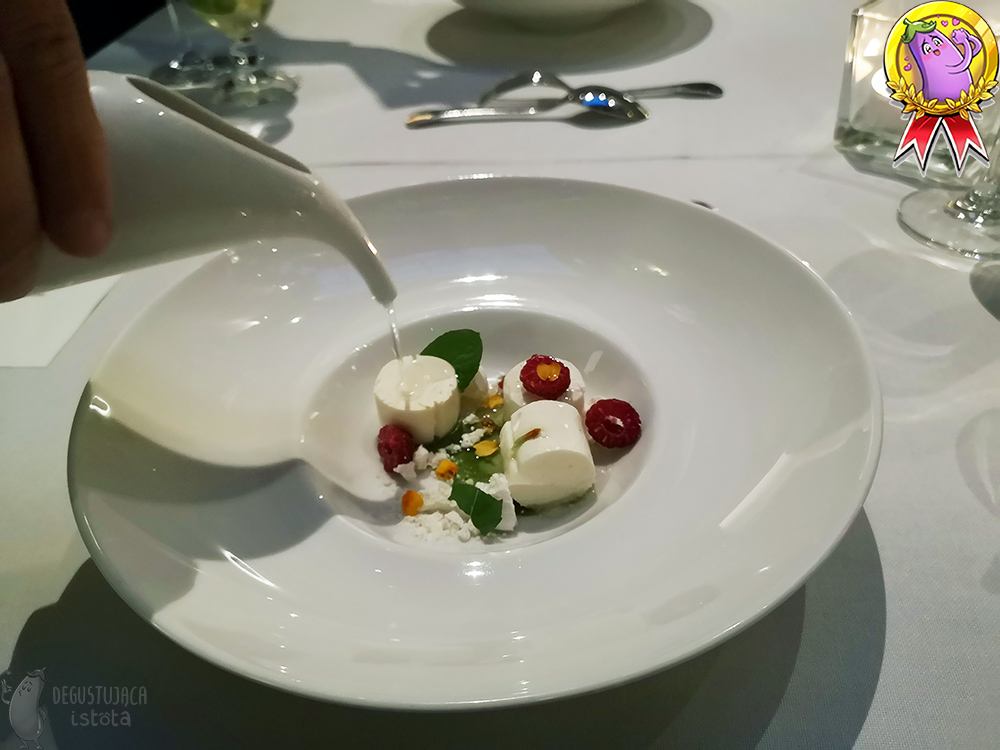On a white plate, white pieces of parfait and crumbled meringue. There are still pieces of raspberries, orange flower petals and green leaves. The waiter pours a clear consommé from a ceramic white pitcher.