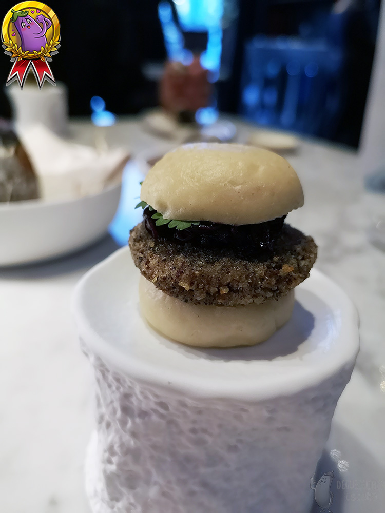 On a small white pedestal there is a burger in a cut-up steamed bun. Underneath the top half of the roll a green coriander leaf sticks out.