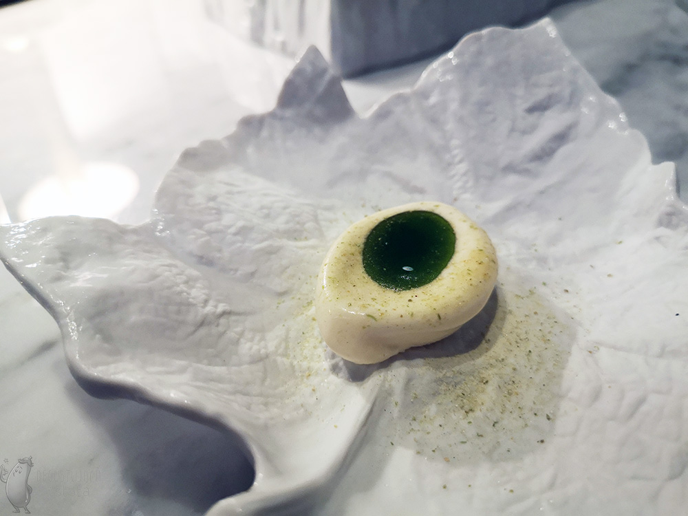 Białe lody z zieloną oliwą nalaną w zagłębienie w lodach, ułożone na białym talerzu w kształcie liścia.