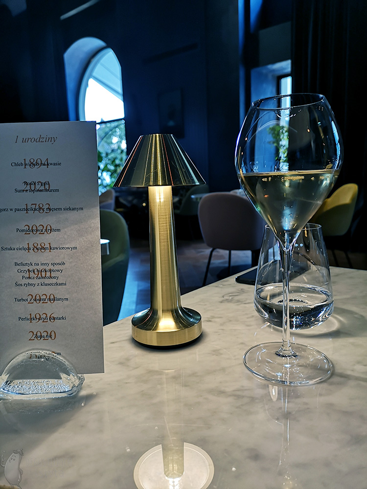 Kieliszek szampana na stole, obok lampka i menu włożone w stojaczek na menu.