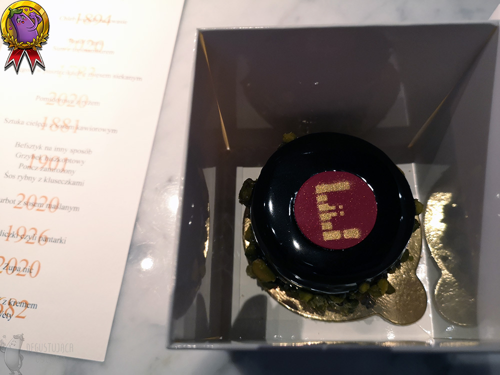 Wnętrze pudełka. Oblany czekoladą torcik na złotej podstawce, ze złotym E na środku tortu napisanym na czerwonym krążku z czekolady.