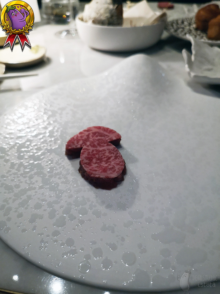 Na płaskim białym talerzu położony jest tatar, przykryty marmurkowym plastrem Wagyu. Mięso ma kształt grzybka.