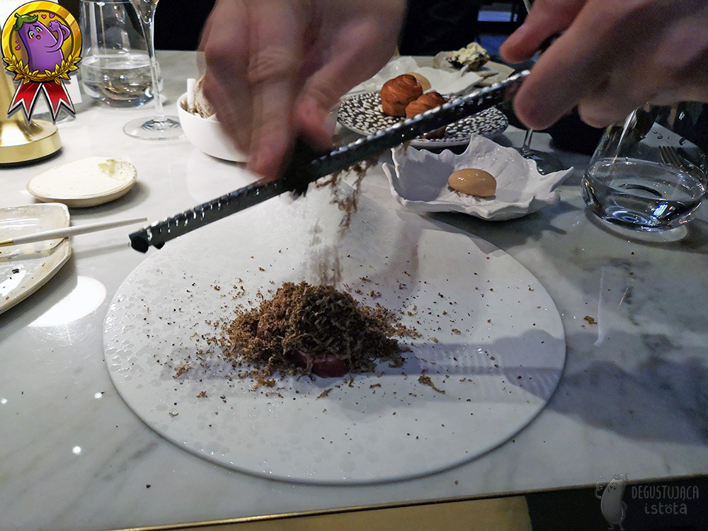 Grzybek z mięsa w trakcie zasypywania go wiórkami tartej przy stoliku trufli. W oddali na stoliku widać lody borowikowe na talerzyku w kształcie białego liścia i bułeczki.