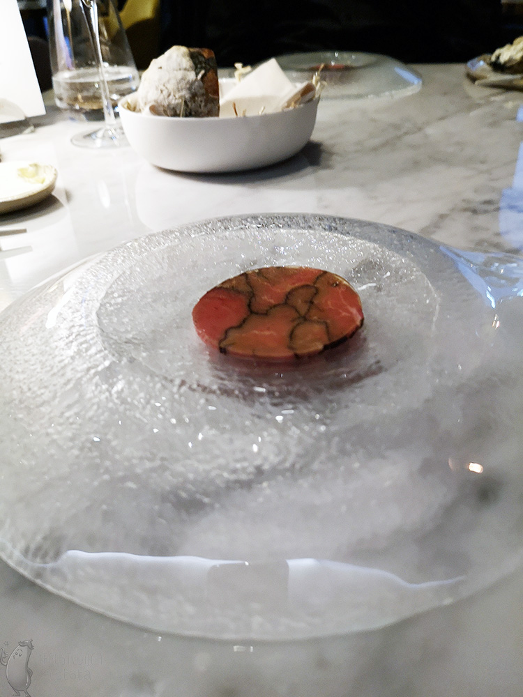 Na dużym, szklanym, lekko przezroczystym talerzu leży okrągły plaster sprasowanego mięsa z czarnymi obrysami w środku.