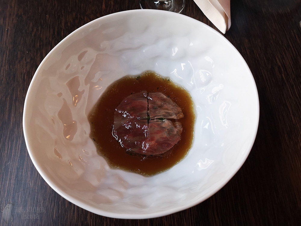 In a deep white plate is a dark broth with a slice of wagyu in the middle.