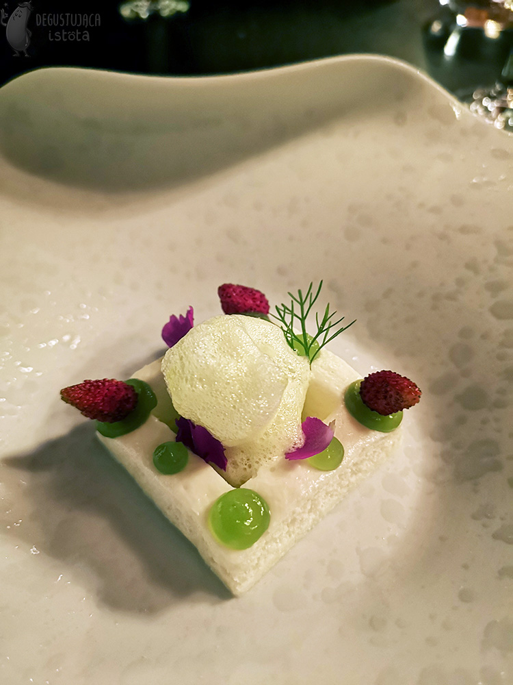  Arranged on a white plate with an uneven surface there is a square shaped white foam with the center cut also into a square. There are green gel dots on the edges of the foam, and three of them contain a wild strawberry. Another white foam is applied inside. The dessert is decorated with several pink petals.