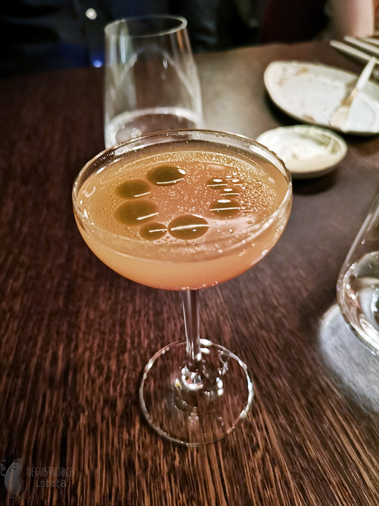 A glass with a rose-orange drink, more transparent at the top and denser at the bottom. Greenish oil circles float on top of the drink.