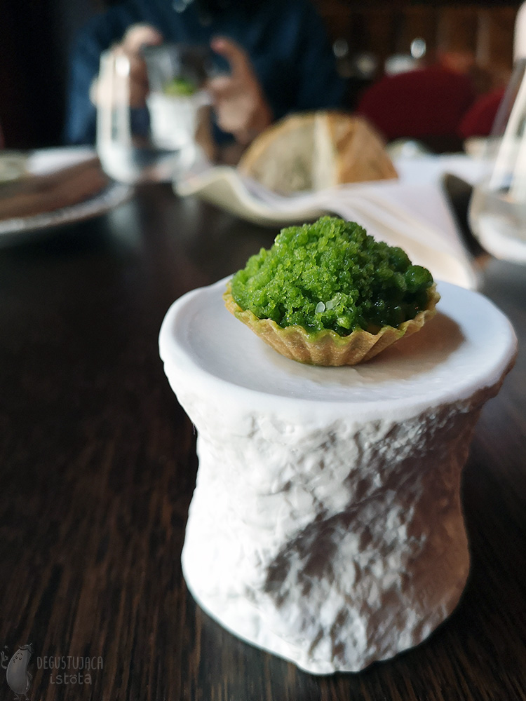 A small tartlet with green granita lies on a white ceramic pedestal.