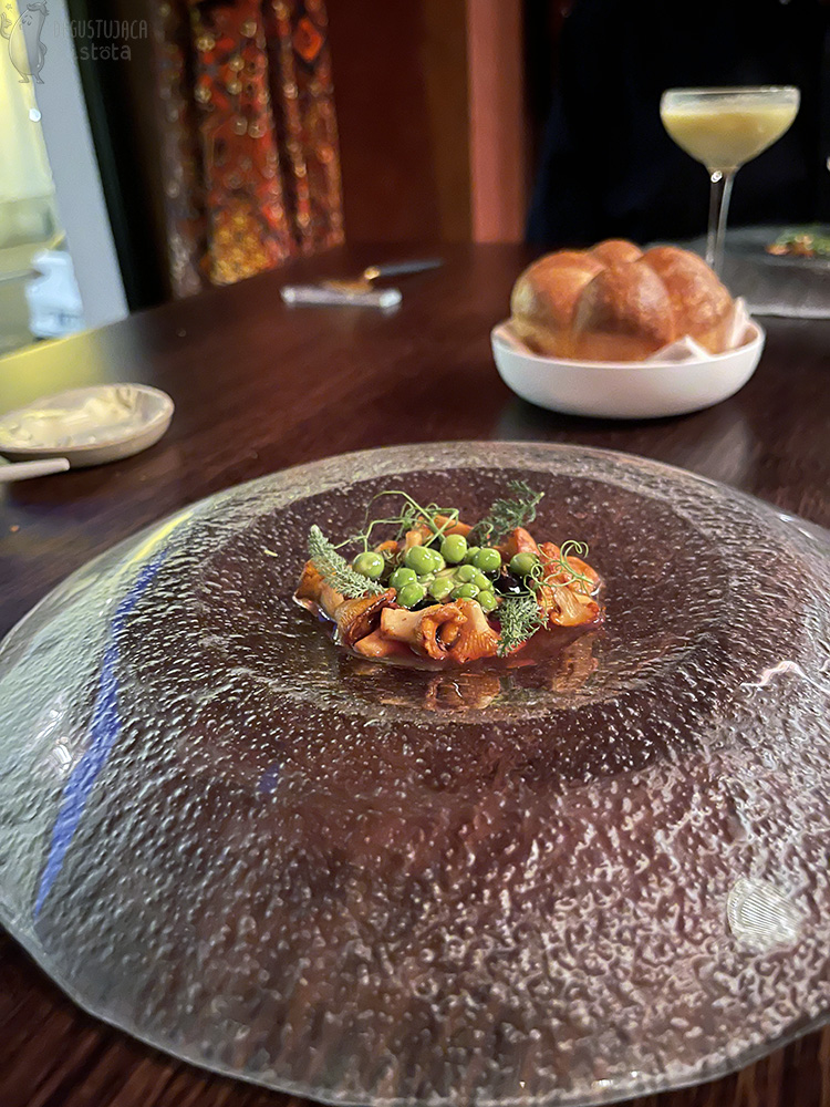 In a large transparent plate, there are chanterelles with sauce and green peas. In the background in the glass you can see a yellow, creamy drink and small brioches.