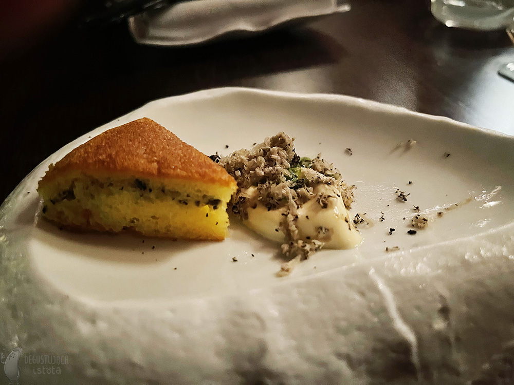 On a white plate, stylized on marble, there is a white ice cream topped with coarse grated black truffle. Next to it, on the same plate, is a triangular piece of potato cake.