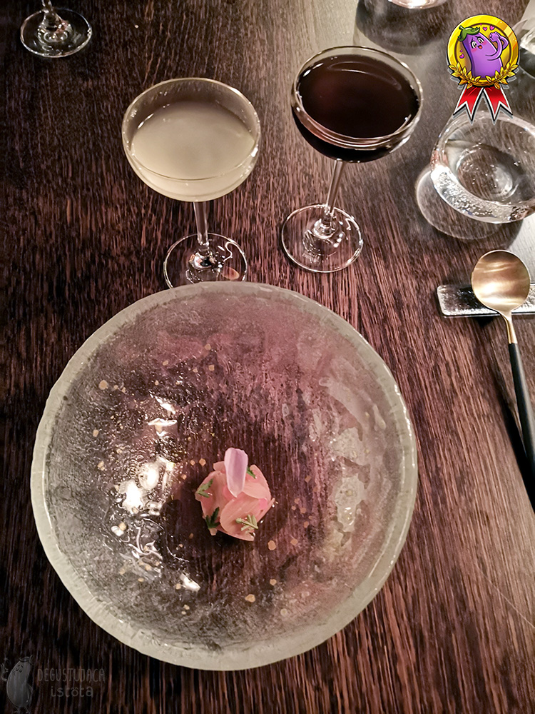 In a deep glass, In a deep transparent glass bowl, pink rhubarb slices with a rose petal on top lie arranged in the middle. Two glasses stand next to it. One with a yellow drink, the other with dark brown rooibos.