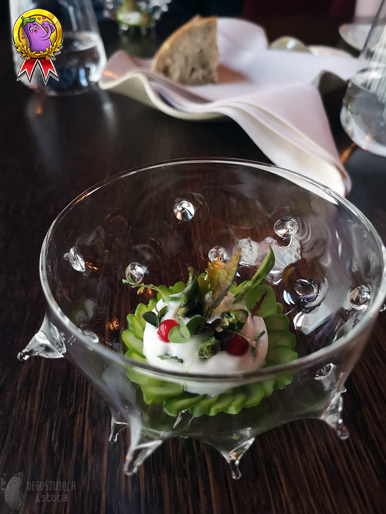 A glass bowl resembling a sea urchin. Arranged slices of green asparagus in the center, on which is a white foam decorated with leaves and two red balls.