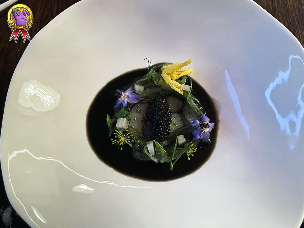 Pieces of white fish are placed in a large, white plate, surrounded by black powder and arranged in the shape of a circle. There is a portion of sturgeon caviar on the fish. There is a black sauce around the fish and it is decorated with purple and yellow flowers and green leaves.