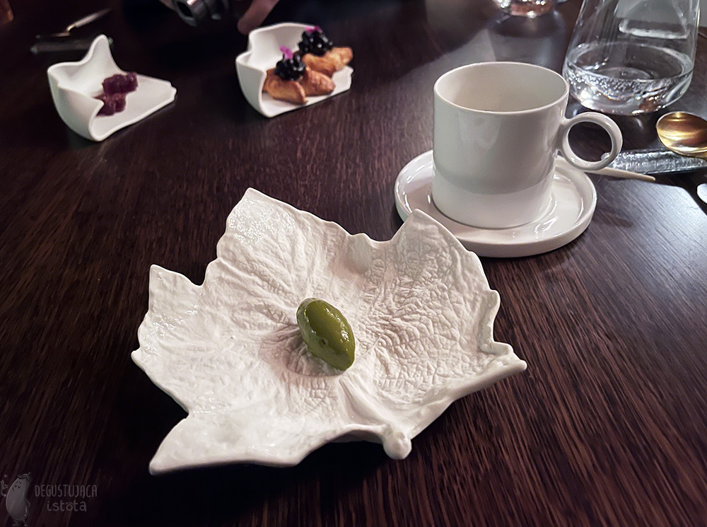 Biały talerz w kształcie liścia klonu na którym jest porcja zielonych lodów. Obok biały kubek, a na dalszym planie dwa małe talerzyki. Jeden z dwoma wypiekami danish z jagodami, drugi z dwoma różowymi galaretkami w cukrze w kształcie kwiatków.