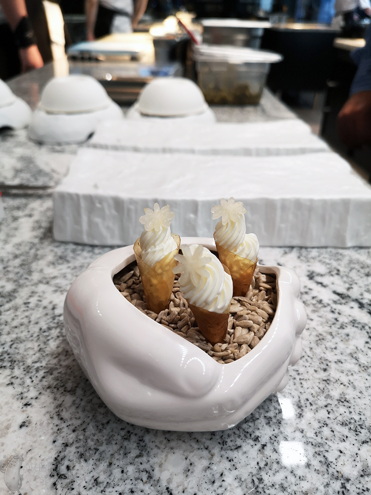 Brązowe rożki wypełnione białymi lodami i ozdobione białym kwiatkiem wyciętym z truskawki. Ułożone są w ziarnach pszenicy w ceramicznej, białej misce w kształcie złożonych rąk.