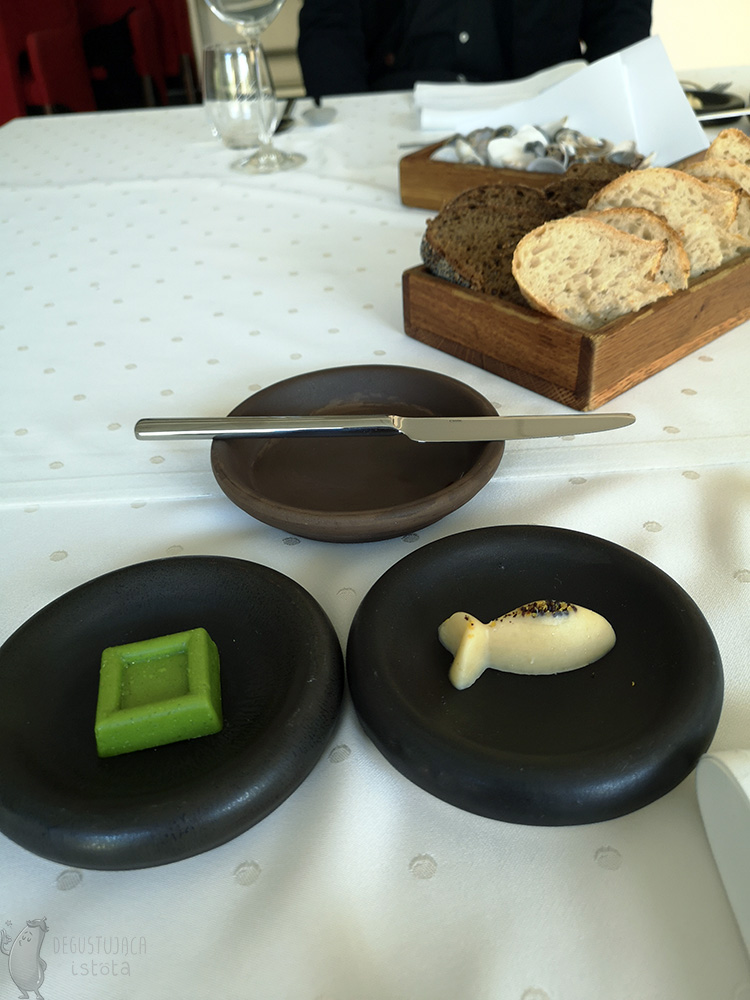 On a table covered with a white tablecloth are 3 black plates and a wooden container with slices of light and dark bread. On one black plate is green butter in the shape of a square and on the second plate is burnt butter in the shape of a fish. On the third plate is a butter knife.