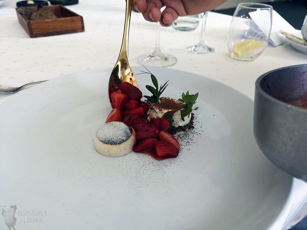 On a large, flat, white plate next to the panna cotta and asparagus ice cream, pieces of strawberries in sauce are placed with a golden spoon.