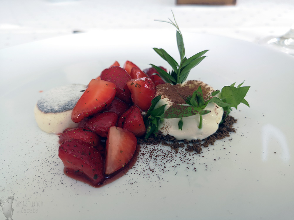 On a white plate lies a portion of white ice cream decorated with green leaves, panna cotta and pieces of strawberries between them.