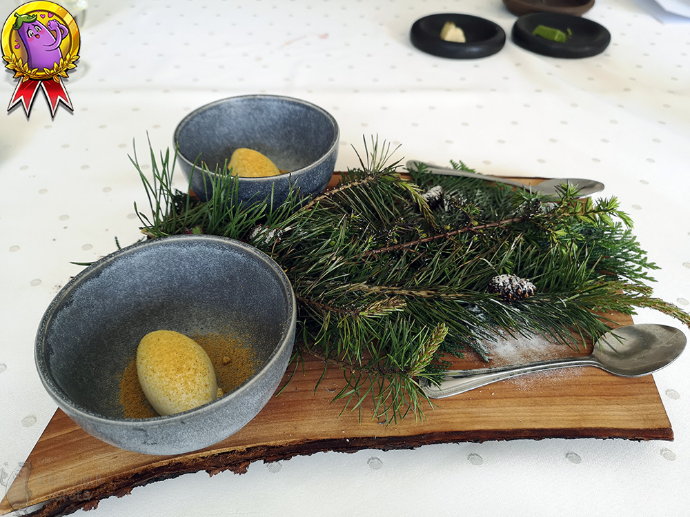 On a table covered with a white tablecloth lies a wooden board on which are two gray bowls with bright ice cream sprinkled with pollen. The board is covered with conifer branches and on them, sprinkled with powdered sugar, are placed small cones.