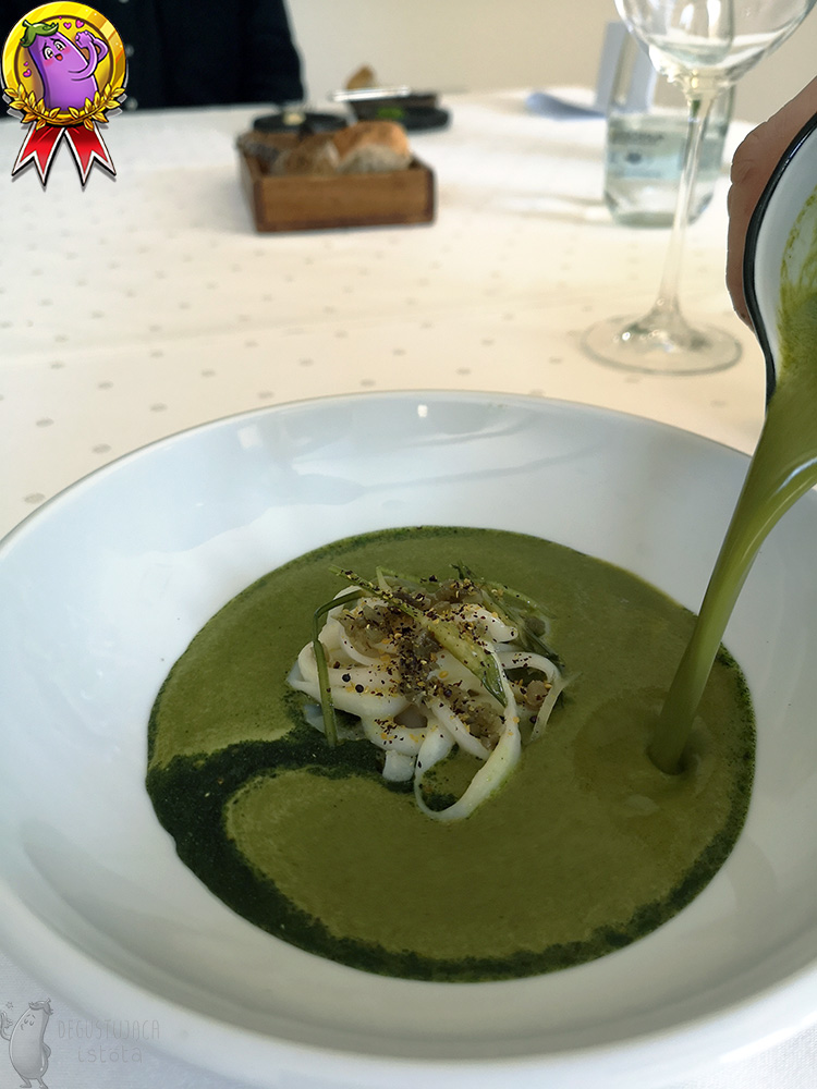 Intensywnie zielona, kremowa zupa w trakcie nalewania do białego, głębokiego talerza w którym znajduje się makaron z kalmara.
