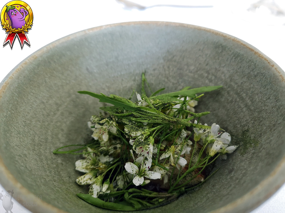 The dark grey bowl is filled with white flowers and stems of cress sprinkled with green powder.