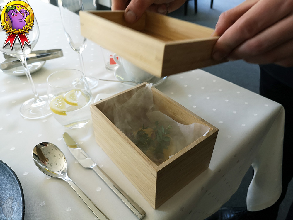 On the table, there is a small box in which there is a tartare with a dried yolk sprinkled on top. Smoke is coming out of the box whose lid has been lifted.