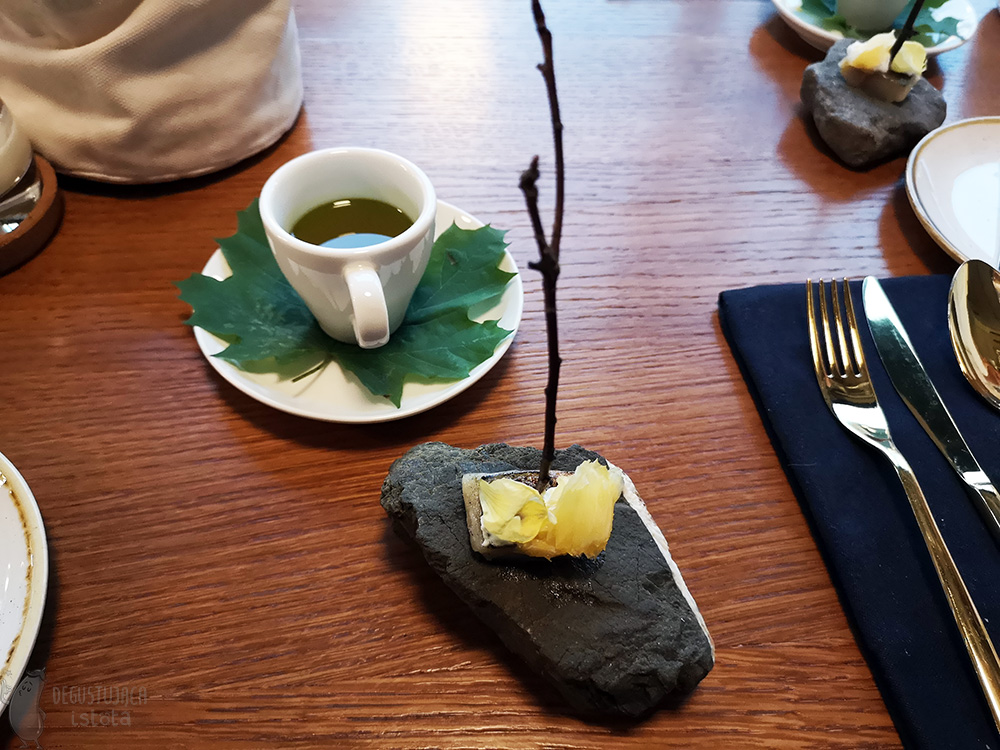 White coffee cup filled with orange and green liquid. On a saucer and under the cup lies a green maple leaf. Next to it on a stone lies a piece of fish pierced with a stick.