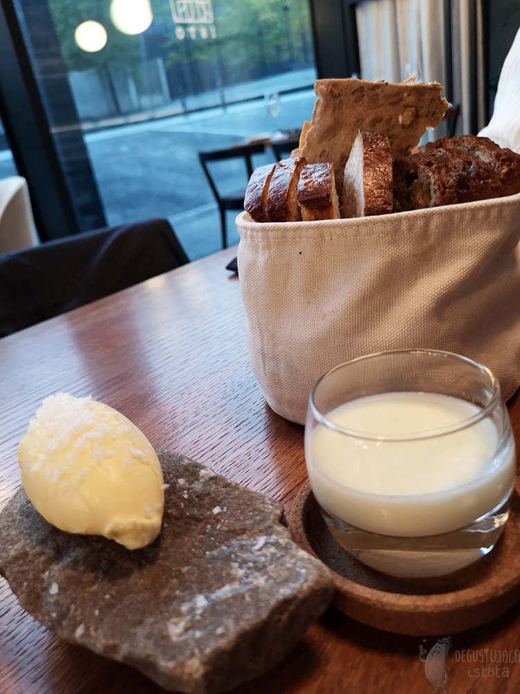 On a wooden table lies a cloth bag filled with bread. Next to it lies a dark stone with a portion of yellow butter sprinkled with coarse salt. In a low glass between the stone and the sack is a white espuma.