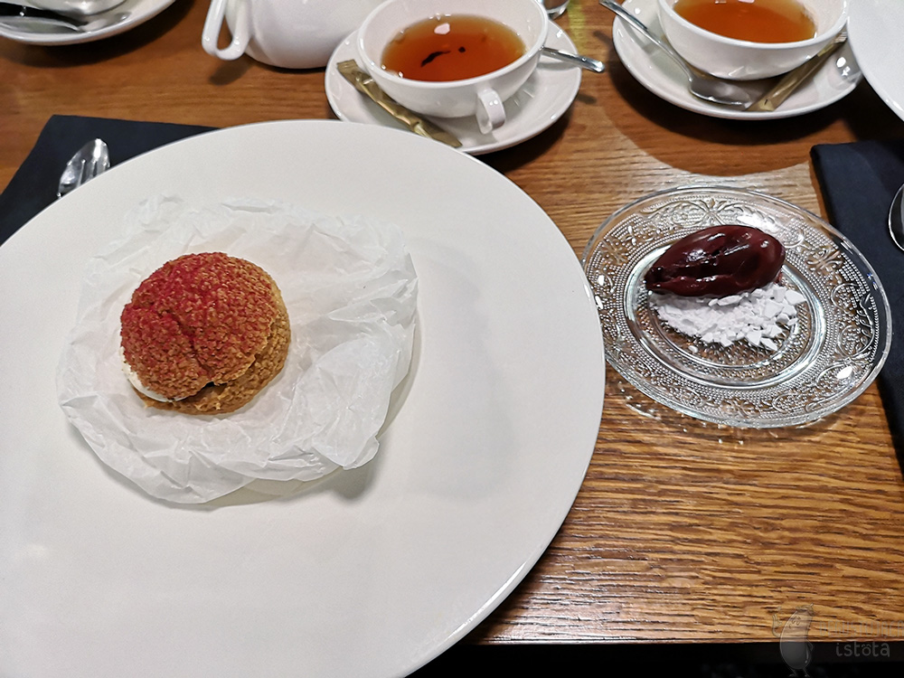 On the table lie white tea cups and a large white flat plate with a puff in the middle. The puff lies on rolled up baking paper. It is sprinkled with red powder on top. On a transparent plate next to the plate with the puff, lies purple red ice cream on crumbled white meringue.