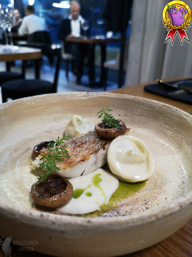 In a gray, flat plate with a high edge lies a piece of white fish with the skin baked up. Next to the fish there are hats of fried red pine mushrooms and two ravioli. The whole dish is covered with a thick white sauce and drizzled with green olive oil.