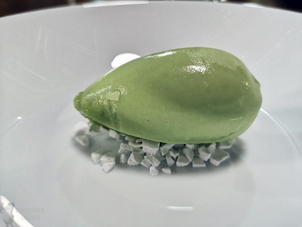 Zielona porcja lodów ułożona na białych okruchach bezy na małym, białym talerzyku.