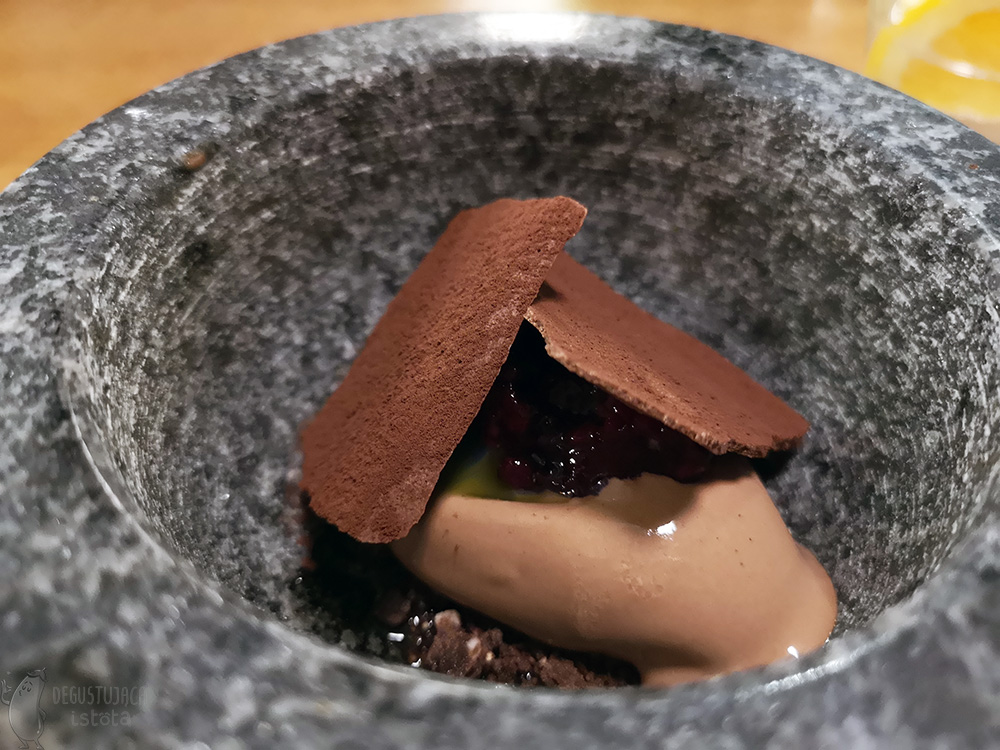 In a dark mortar lie sheets of chocolate and under them you can see a portion of chocolate ice cream and raspberry jam laid on top of the ice cream. The chocolate sheets are dusted with cocoa.
