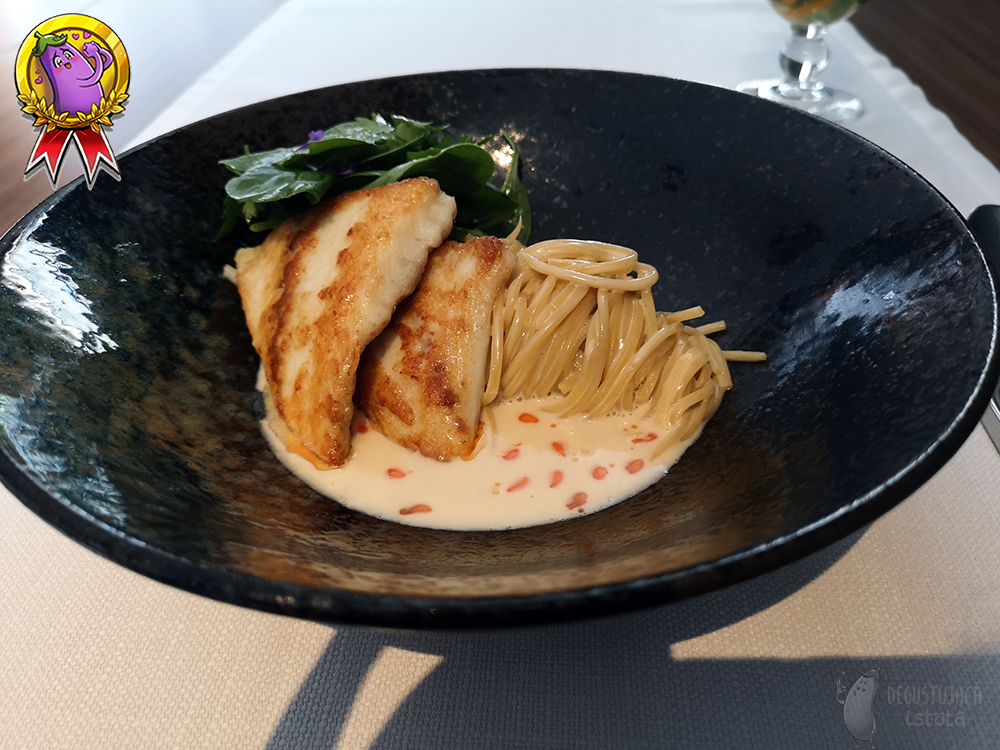 In a black shiny plate lies a coiled linguini. There are two pieces of turbot and baby spinach leaves on the pasta.