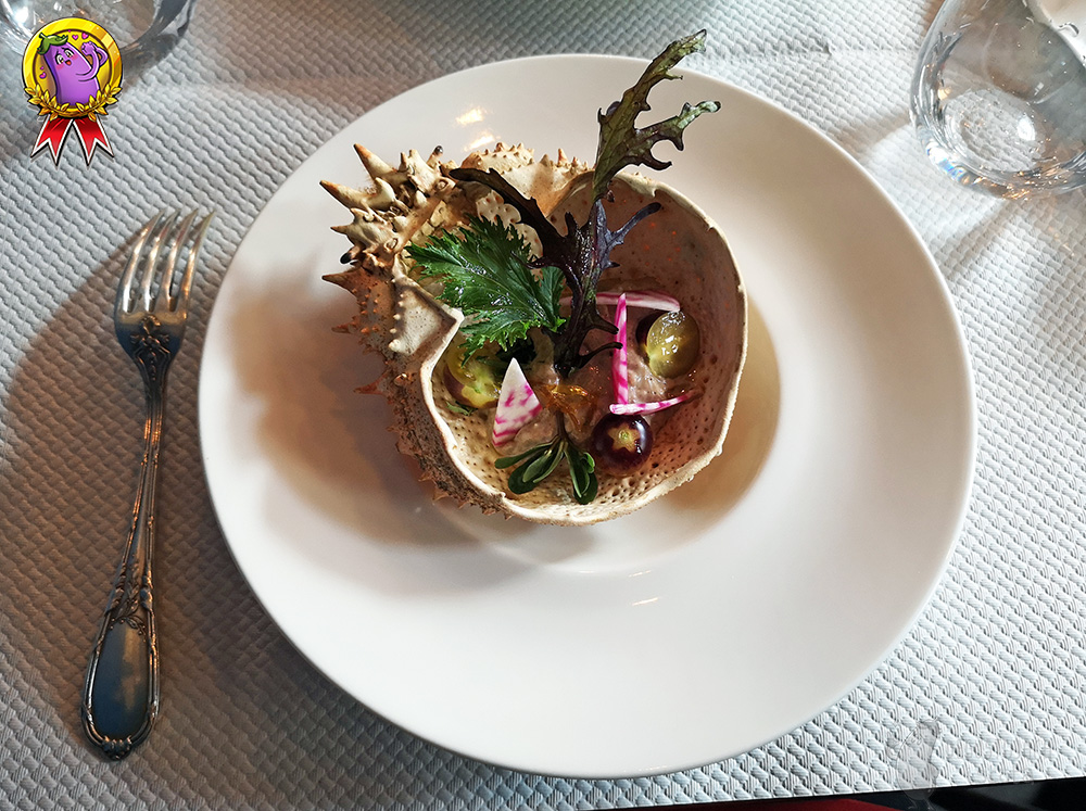 A similar crab shell as before, also on a flat white plate. Inside the shell you can see sliced in half, green and purple small tomatoes, lettuces, pink and white beet sticks and gray mousse. Next to the plate lies a beautifully decorated silver fork.