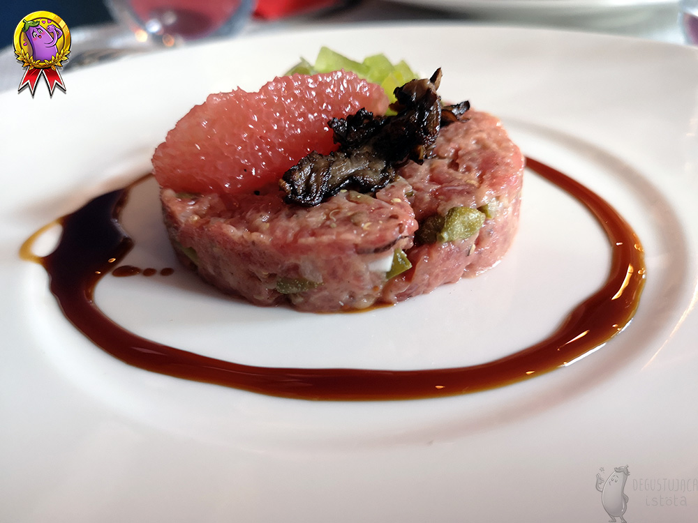 A portion of beef tartare, finely chopped arranged in the shape of a disc. A dark sauce is poured around it and the top of the tartare is garnished with a grapefruit fillet and slices of dark mushrooms.