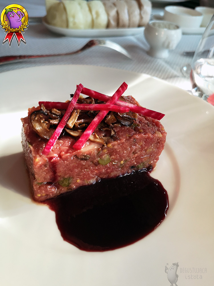 A huge portion of finely chopped beef tartare, arranged in a cuboid on a white plate. Slices of mushrooms are arranged on top of the tartare and pink and white beet posts on top. A portion of dark brown sauce is placed next to the tartare.