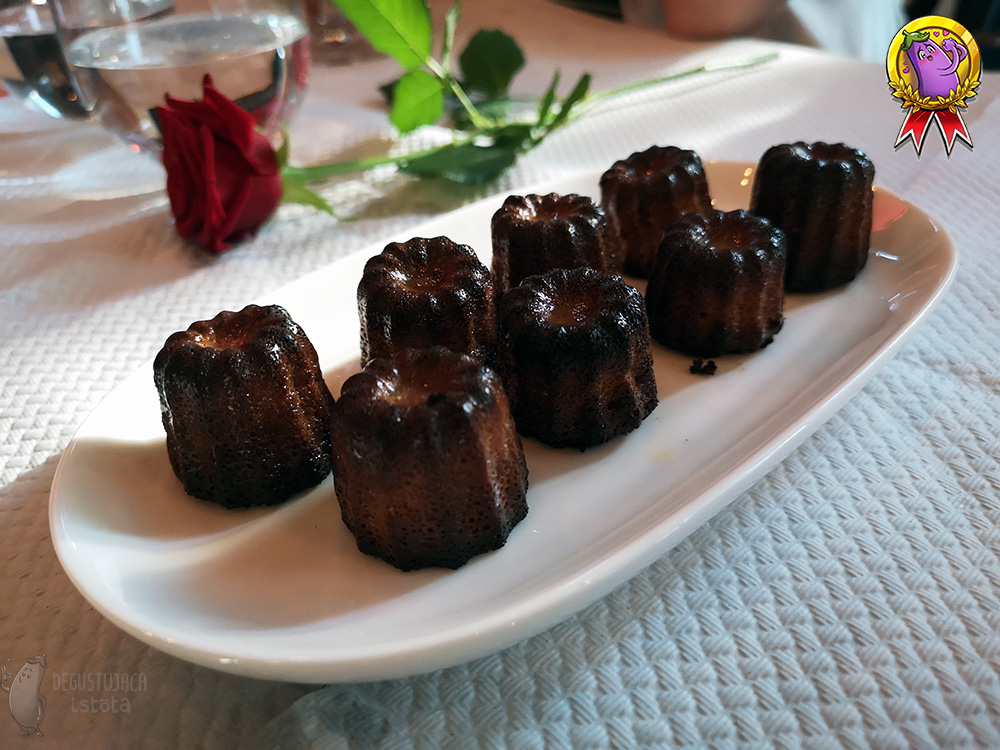 Eight Cannelé are arranged on a white oval plate. They are dark brown. In the background behind the Cannelé lies a red rose.