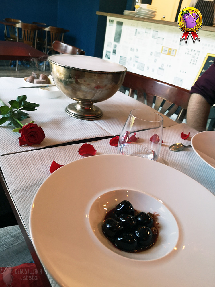 White large plate with a deep center in which are large, black, shiny cherries in caramel. In the background, on a table covered with a paper white tablecloth you can see a silver bowl with still floating steam. Next to the bowl lies a red rose. Around it, rose petals lie on the tablecloth.