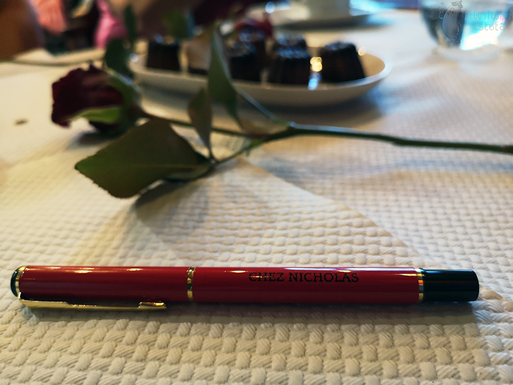 Czerwono złoty długopis ze złotym napisem Chez Nicholas leży na stole a za nim w tle czerwona róża i talerzyk z canele.