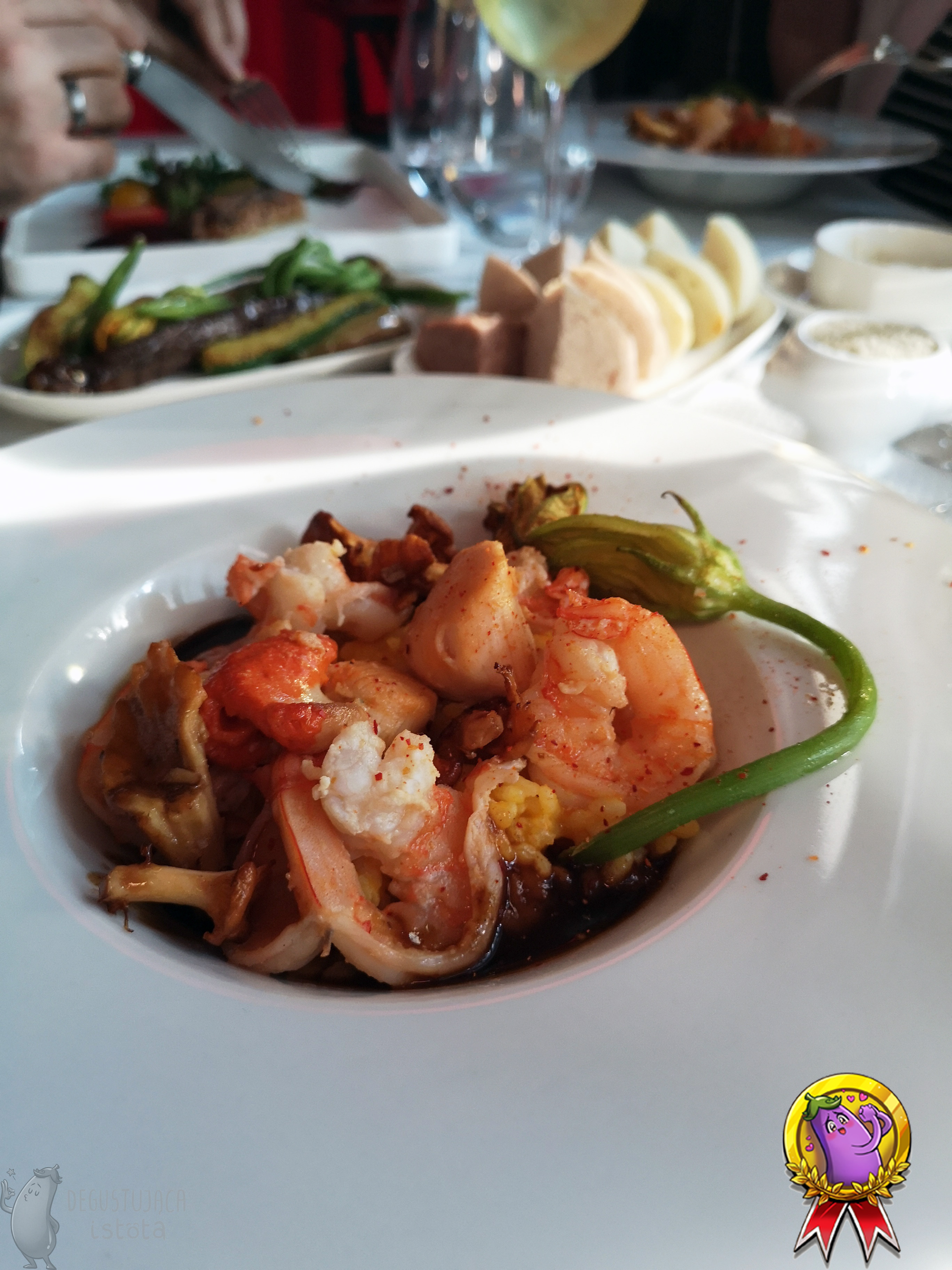 A white plate full of shrimp, scallops and chanterelles. A hint of yellow rice can be seen underneath. A steamed zucchini flower is placed on top of the seafood.