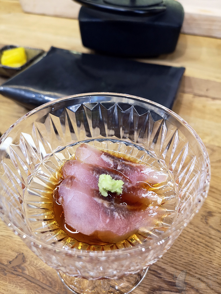 In a transparent dish are arranged three thin slices of raw fish. They are doused in dark sauce and garnished on top, with a small portion of grated green wasabi.