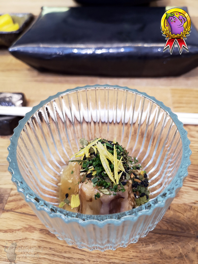 In a transparent container there is a piece of eggplant peeled and garnished with spring onions and strips of lemon zest.