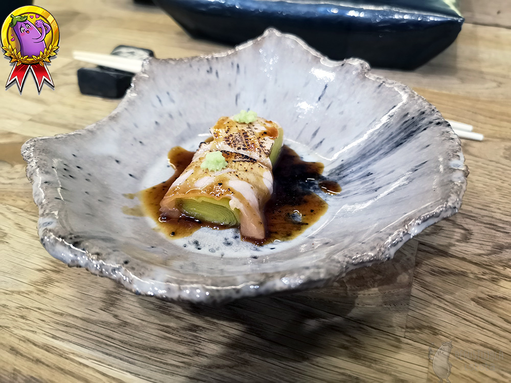 In a light beige ceramic bowl, two pieces of leek are placed in, which are covered by slices of baked salmon. There is a portion of wasabi on the salmon slices. The whole dish is sprinkled with dark sauce.