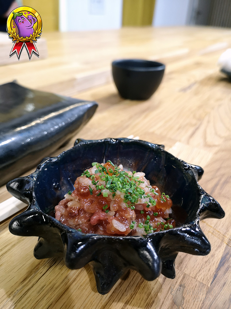 W ciemnej miseczce, stylizowanej na muszlę jeżowca, znajduje się porcja różowego risotto udekorowanego sporą ilością szczypiorku.