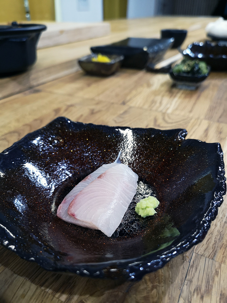Dwa cienkie plasterki jasnej ryby położone w ciemno-brązowej miseczce. Obok ryby leży porcja wasabi.