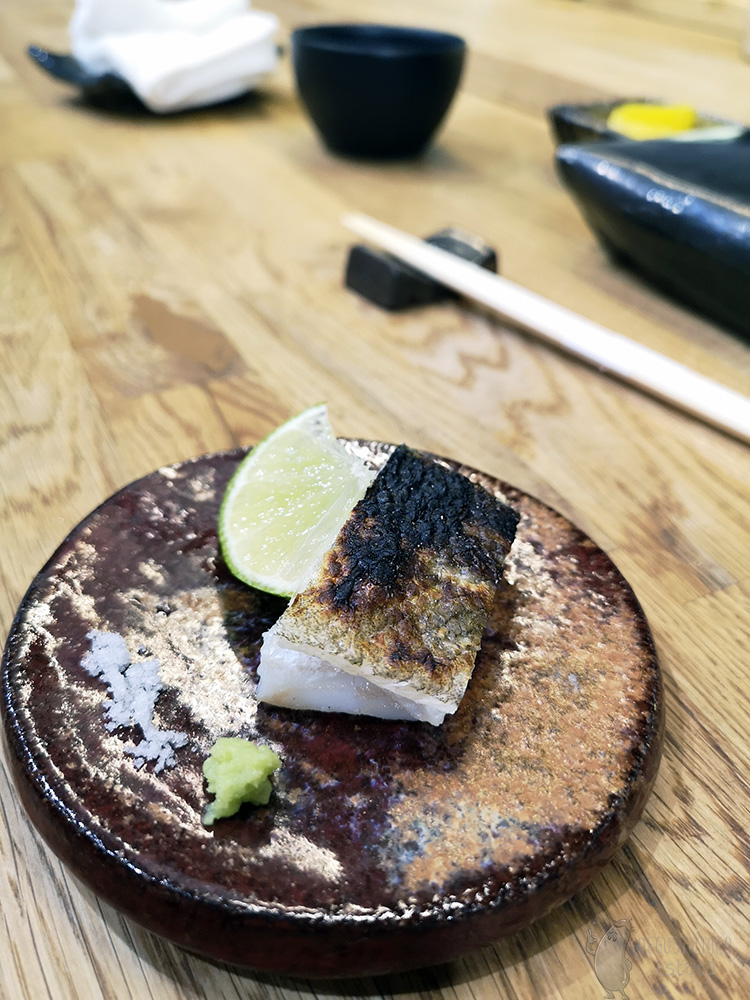 On a dark brown, small plate is a piece of fish with burnt skin. Next to it lies a slice of lime, some salt and grated wasabi.